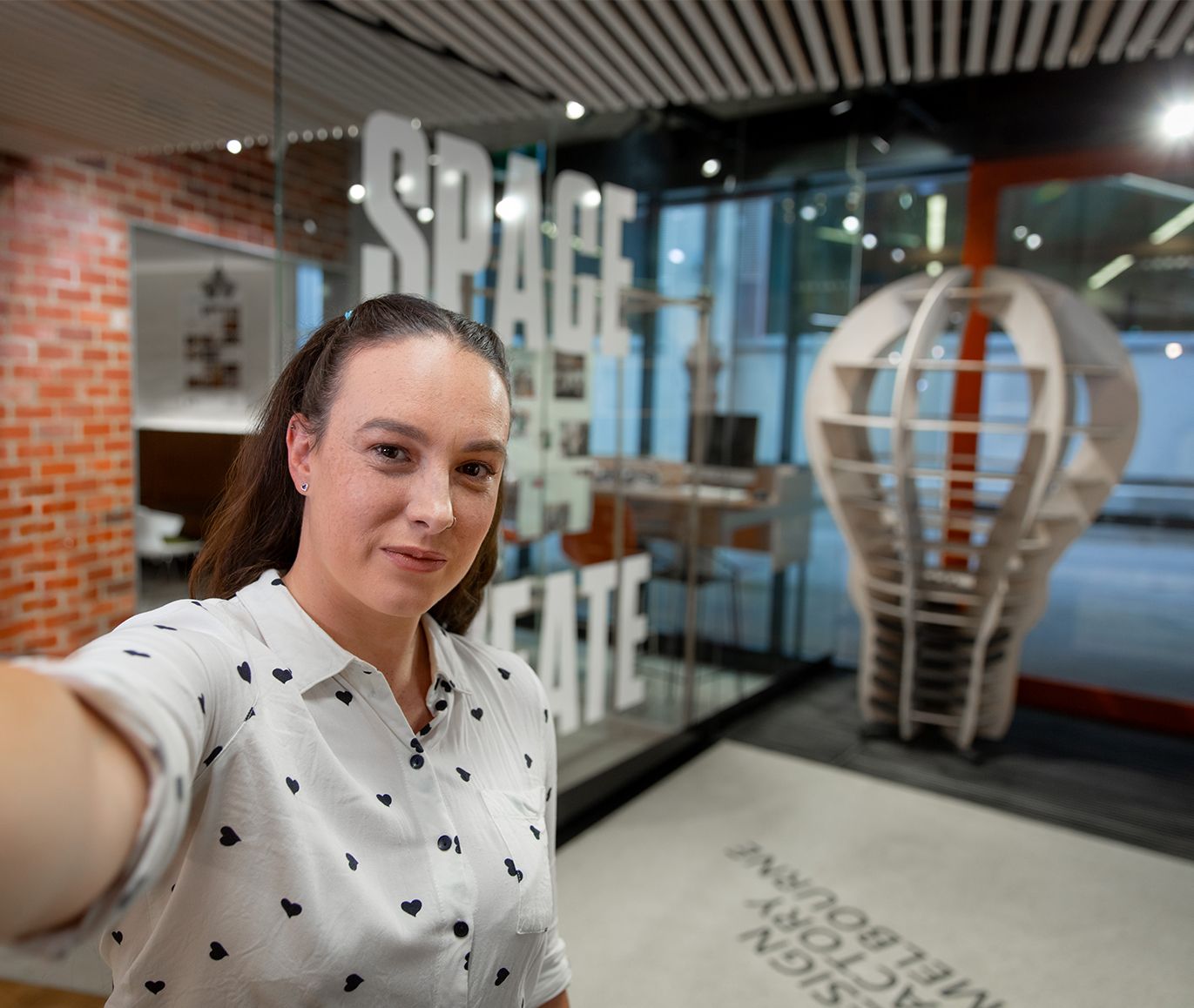 Female student takes a photo in a selfie style shot, smiling at the camera with a Swinburne Hawthorn campus space seen in the background.