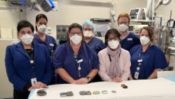Image of seven people in surgical masks and scrubs standing behind a hospital bed looking at the camera, surrounded by surgical equipment.