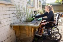A woman in a wheelchair in her backyard gardening with a raised garden bed.