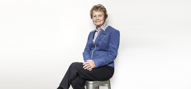 An older woman dressed in a blue jacket and black pants sits in a relaxed pose on a stool