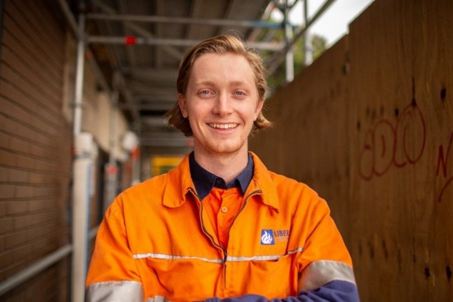 A smiling young man wearing an orange high-vis work shirt stands in front of a construction site