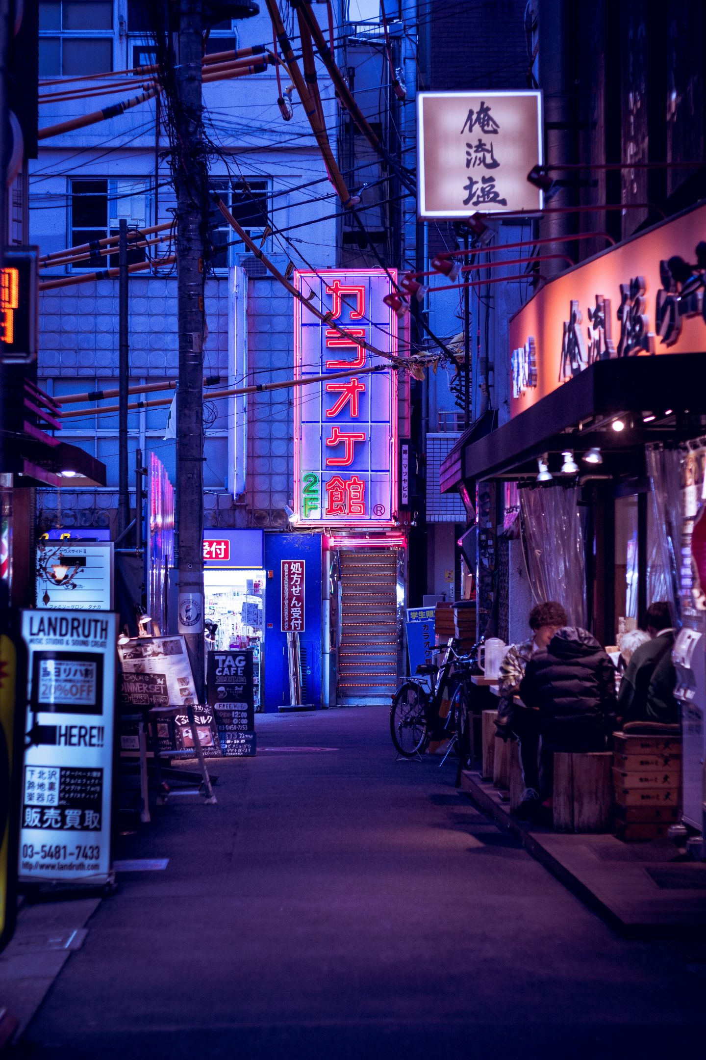 A night scene of a Tokyo side street featuring neon signs, bicycles and people enjoying food in the street
