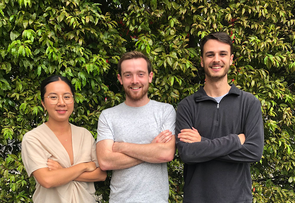 Three students stand in height ascending order from left to write. The young woman on the left is wearing a cream coloured top and clear glasses, she is folding her arms. The young man in the middle wears a grey t-shirt and is smiling at the camera. The young man on the right wears a dark jumper, he is also smiling and folding his arms.
