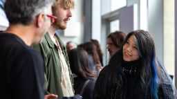 A young woman with long dark hair with blue streaks, wearing a black scarf, smiling in conversation with two men at the Swinburne Study Expo, with other attendees visible in the background. 