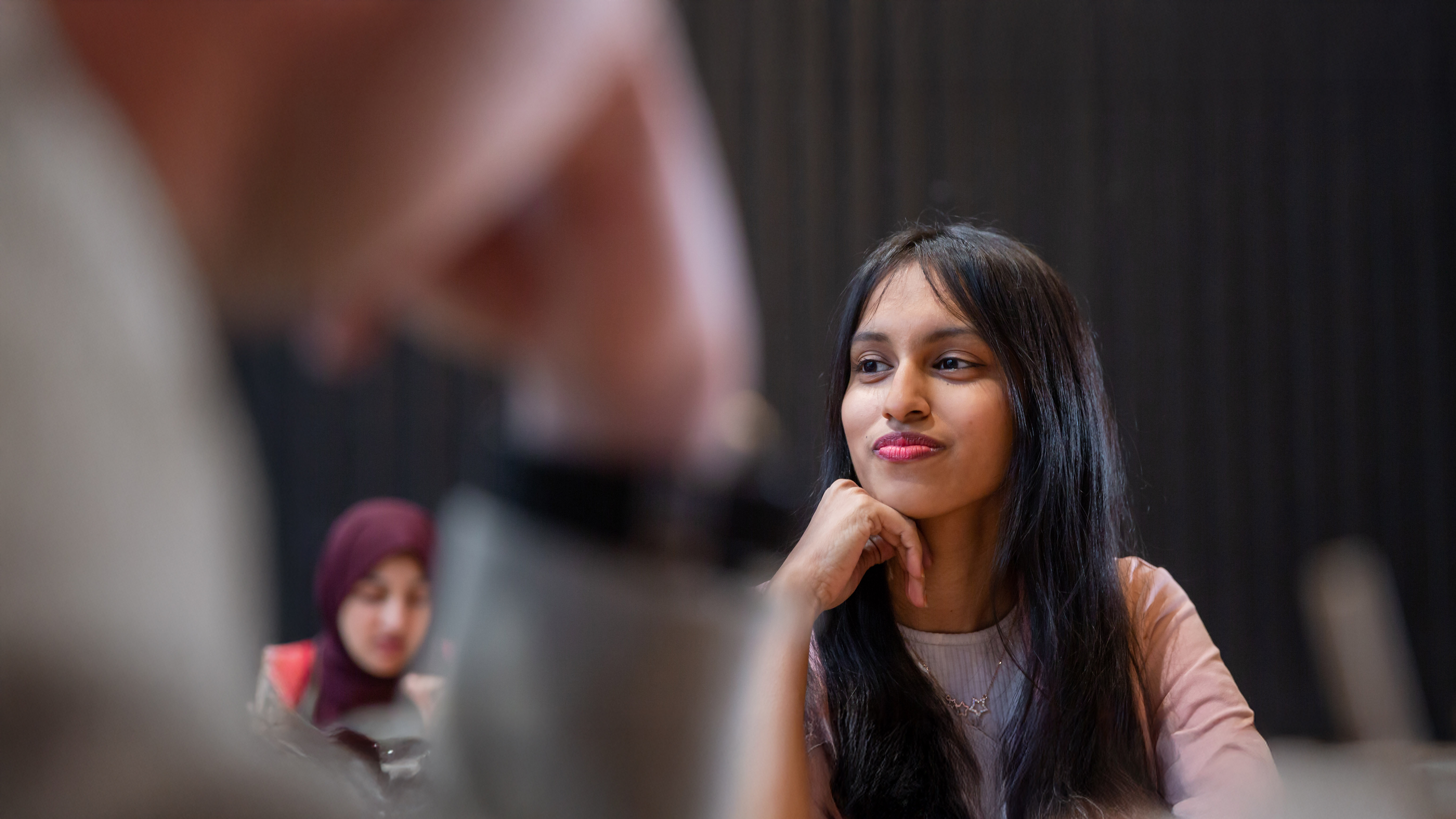 A student with long dark hair sits at a table indoors, resting her chin on her hand and listening attentively, with a takeaway coffee cup in front of her and other students blurred in the background.