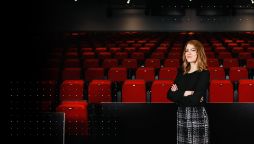 Female student stands alone in a dimly lit lecture theatre,  a sea of red chairs make up the background. In the foreground Natalie stands with her arms crossed and in smart casual clothes.