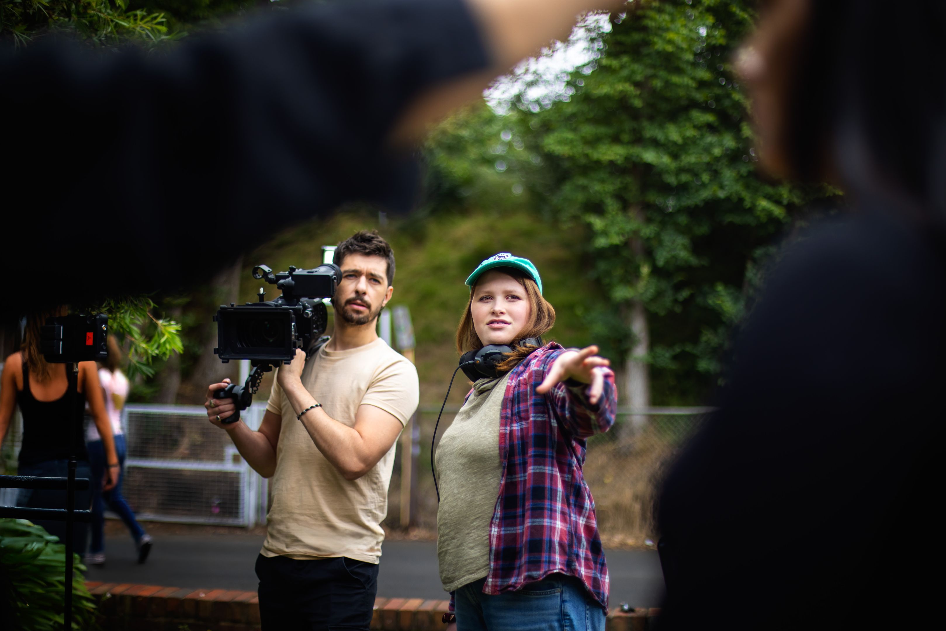 A film crew works outdoors, with one person operating a professional camera and another giving directional guidance in front of a leafy backdrop.