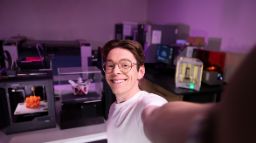 Male student smiling at the camera taking a selfie style photo, in the background shows a dimly lit science lab. 
