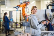 A young woman using a handheld controller in a robotics lab, with others working near a large industrial robot in the background.