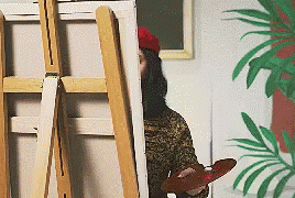 Man leans out from behind an easel to get perspective on what he is painting. 