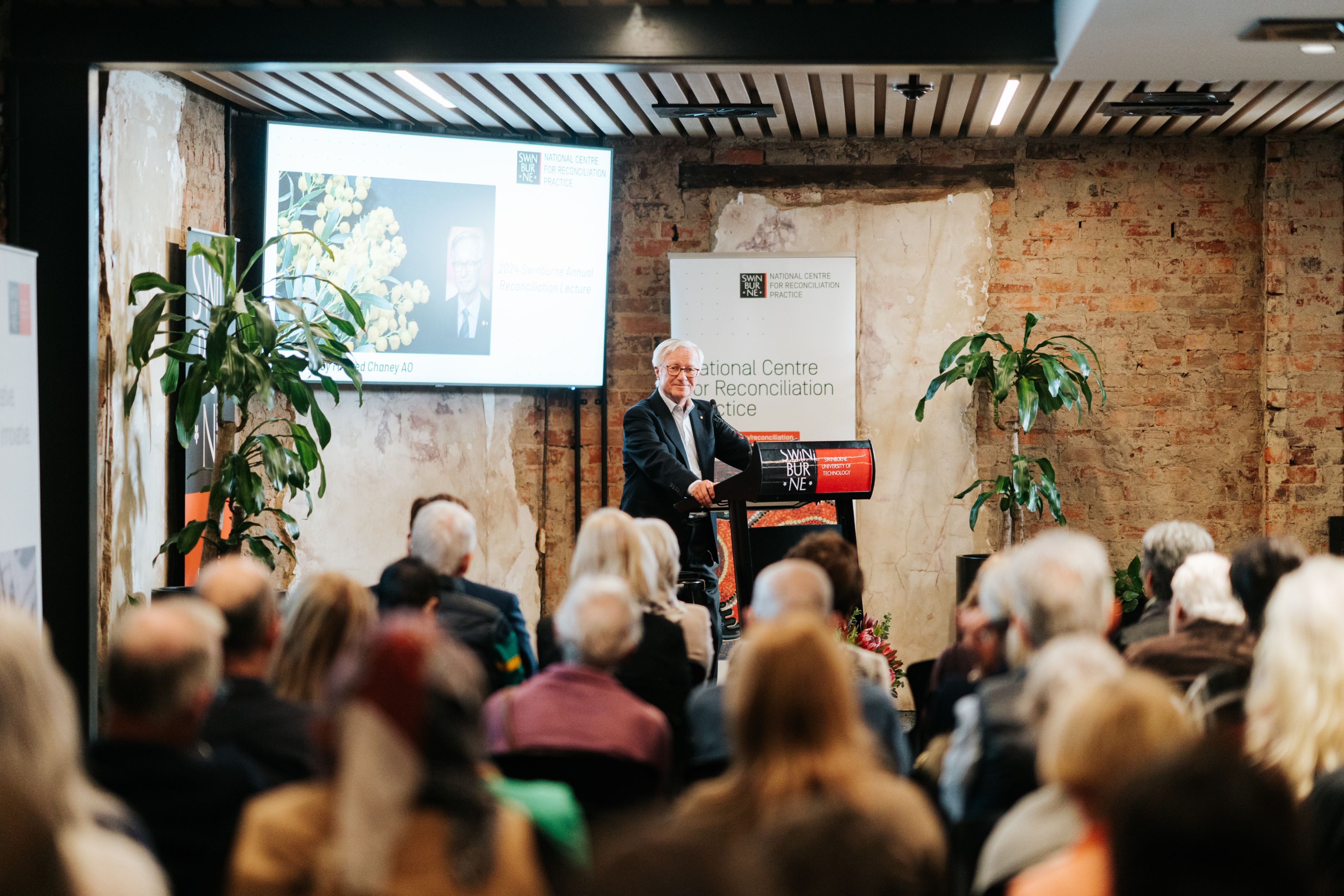 A man delivers a lecture at a lecturn in front of a seated crowd