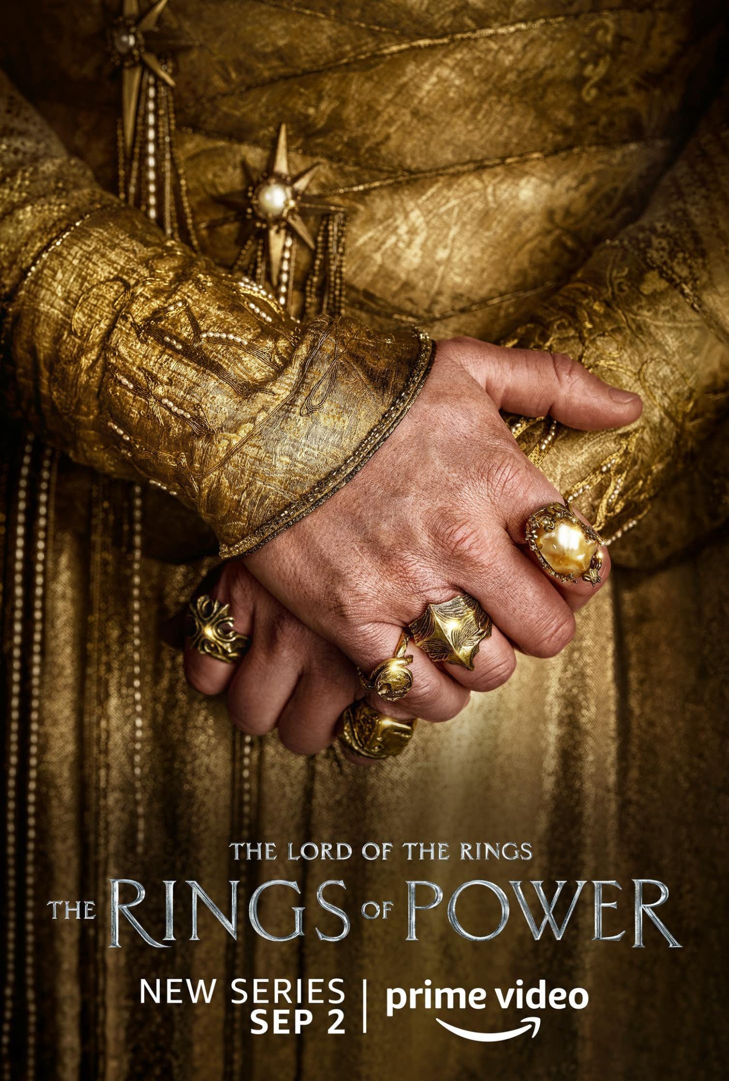 A poster of Amazon Studios upcoming The Rings of Power. someone dressed in gold is folding their hands over with lots of rings visible on their hands