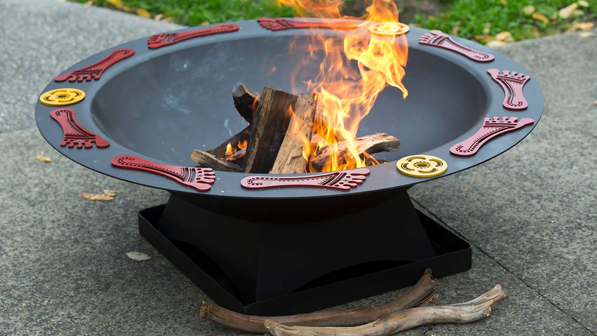 A round, black fire pit with decorative red feet and yellow circle symbols around the edge and a fire burning inside.