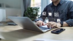 Man typing on a laptop at a desk with icons popping out of the screen related to education, playing video content and online learning. 
