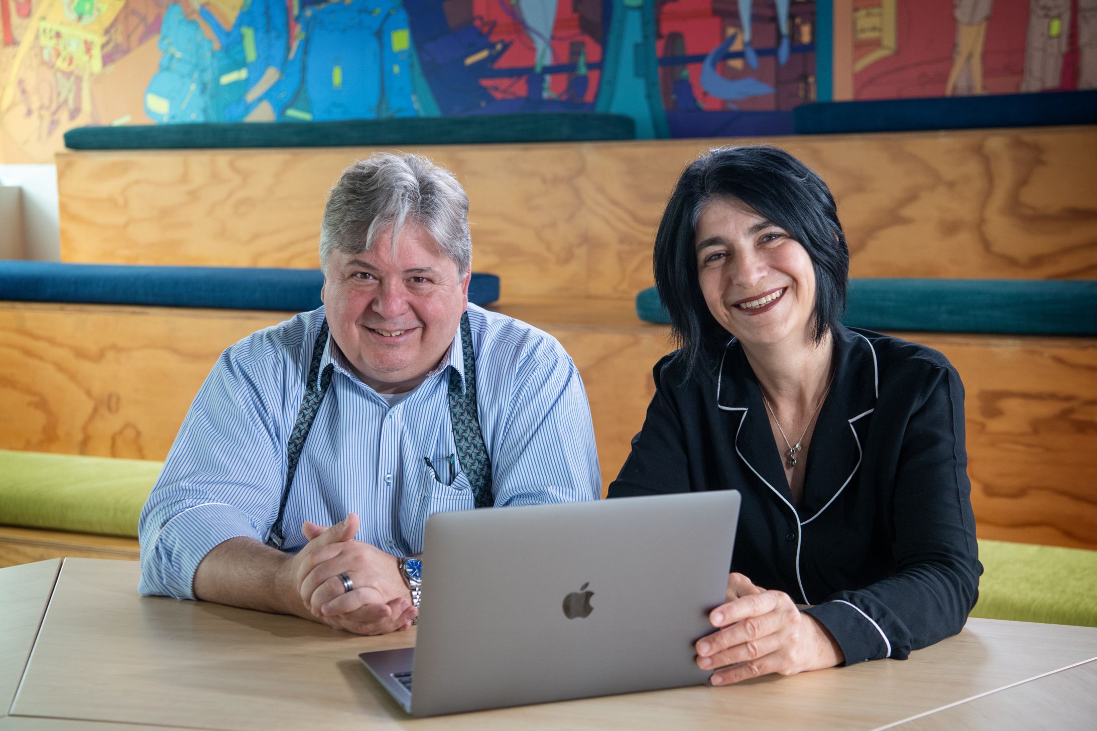 Christopher pictured left in a blue button up shirt and smiling. Gabrielle in a long-sleeve black top smiling with a Mac laptop in front of her on a table