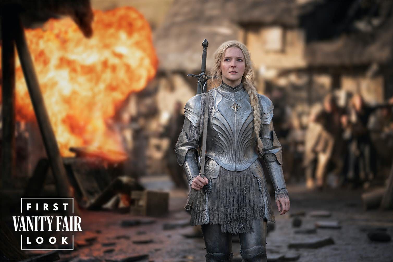 Galadriel played by Morfydd Clark in Amazon Studios Lord of the Rings prequel series is standing armoured on a dirty street in front of a house engulfed in flames
