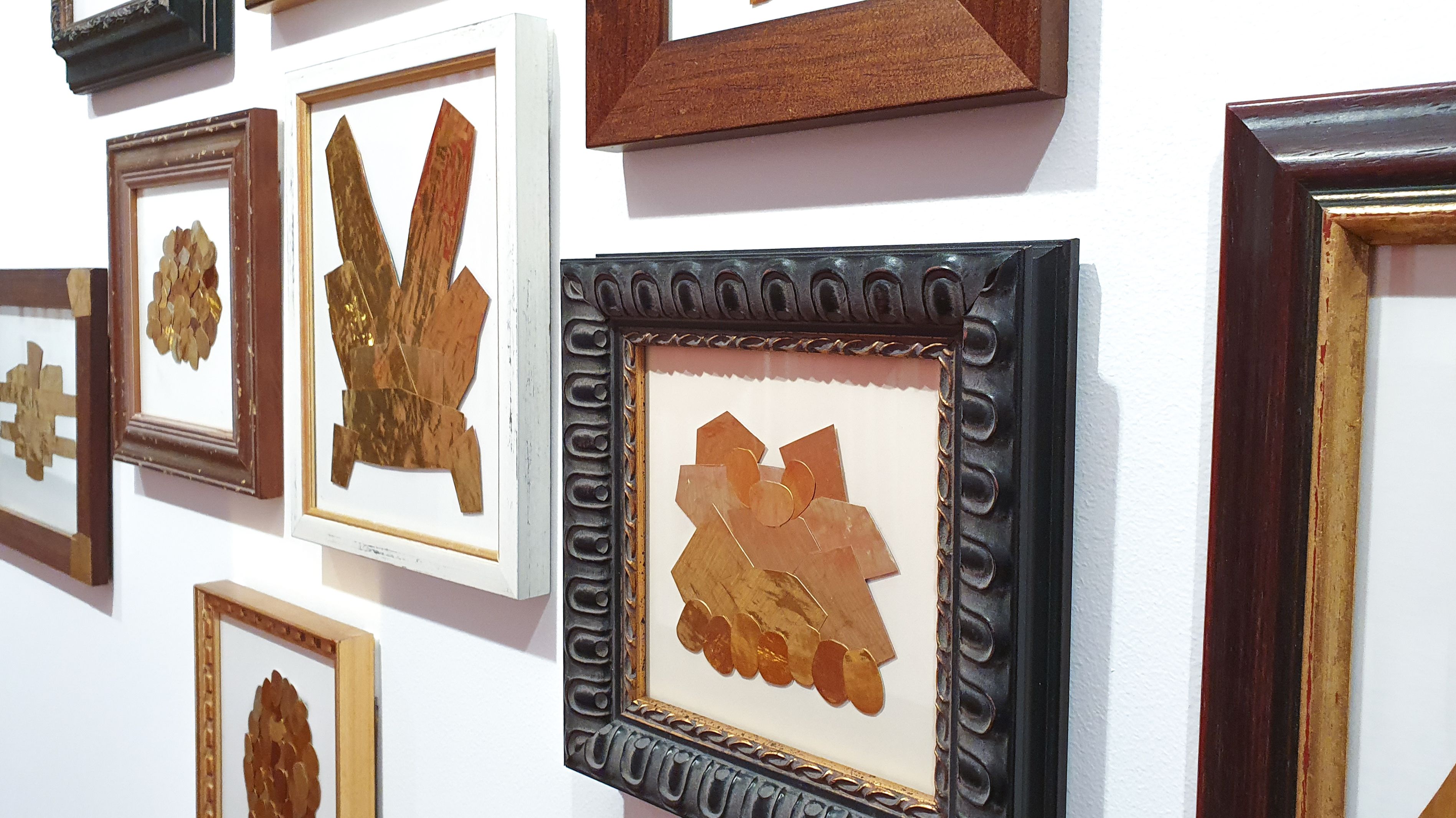 Geometric shapes made from pieces of gold-coloured metal are displayed in an eclectic assortment of wooden frames and hung on a white wall in the Boroondara Town Hall Gallery