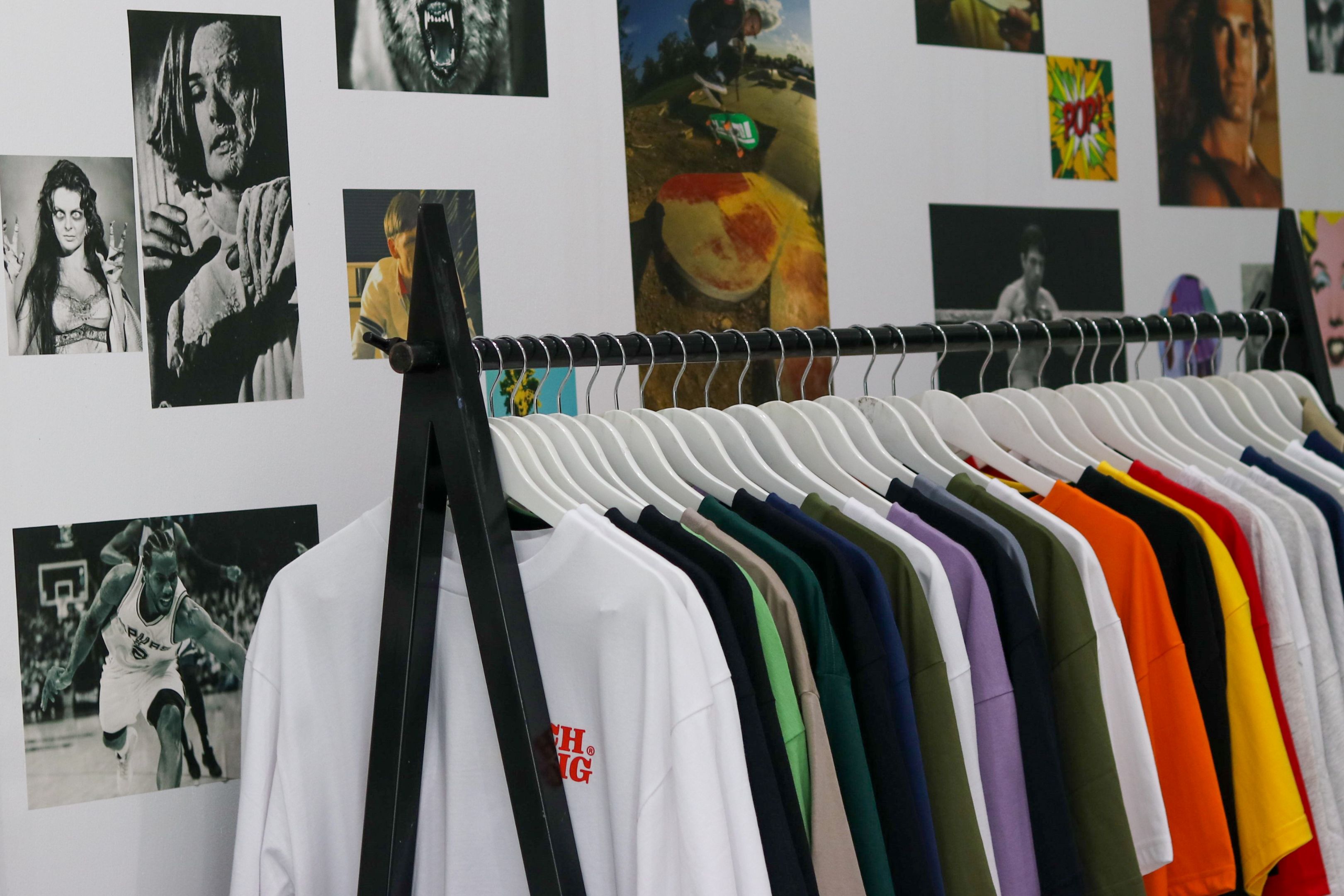 A rack of colorful t-shirts hands in front of a wall covered in photos of skaters, athletes, and iconic pop culture figures 