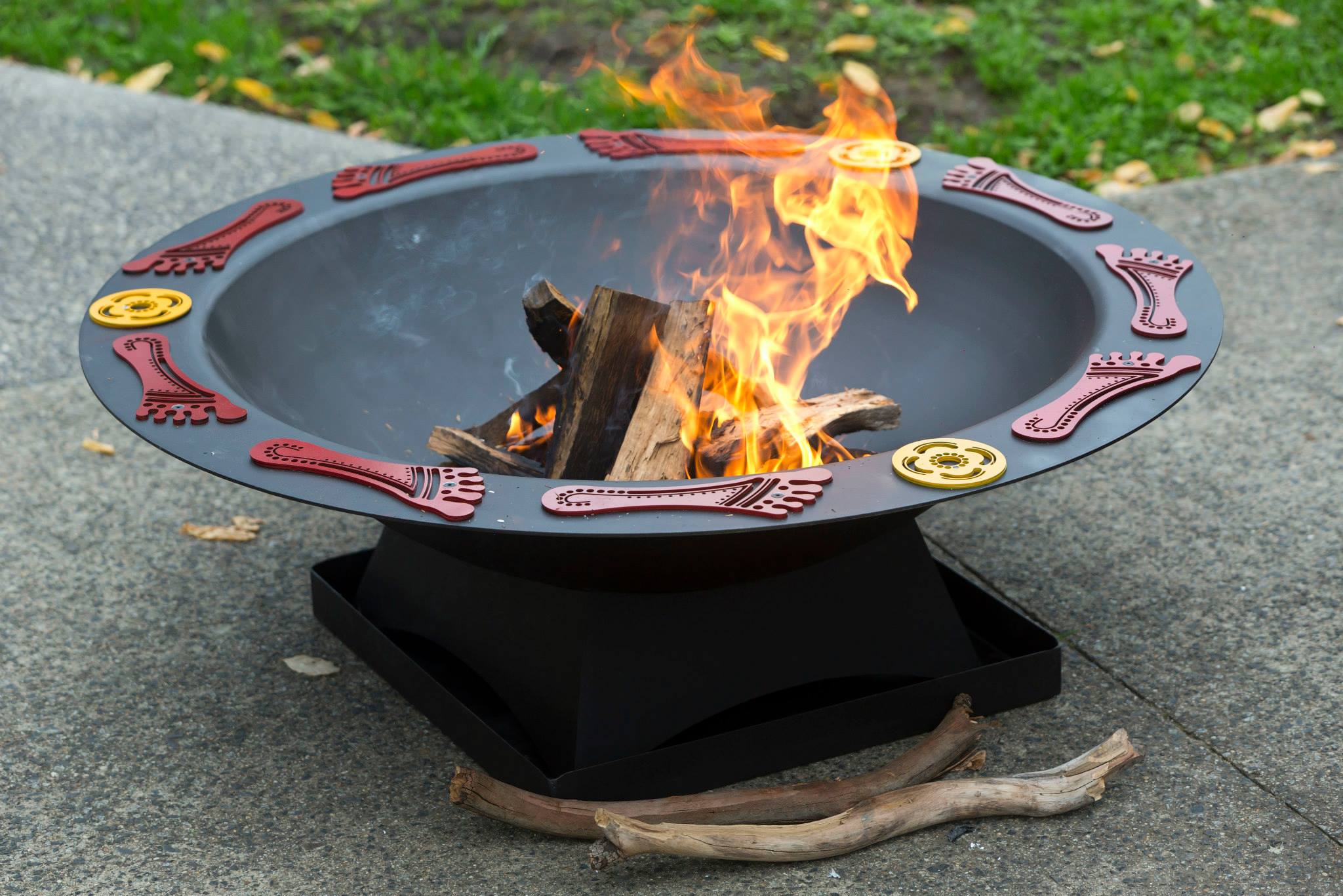 A round, black fire pit with decorative red feet and yellow circle symbols around the edge and a fire burning inside.