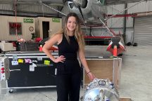 Woman stands in an airline hanger, with a small plane behind her and plane engine beside her.