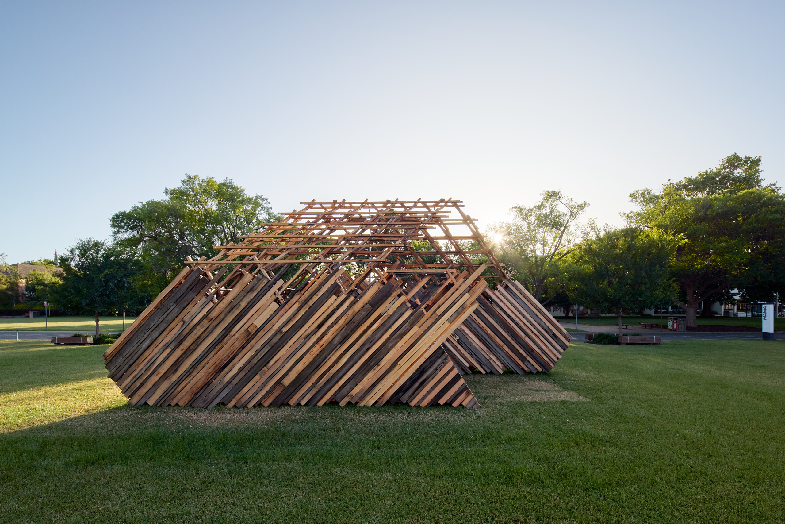A large a-symmetrical structure made from different coloured planks of  recycled wood stands in a green park