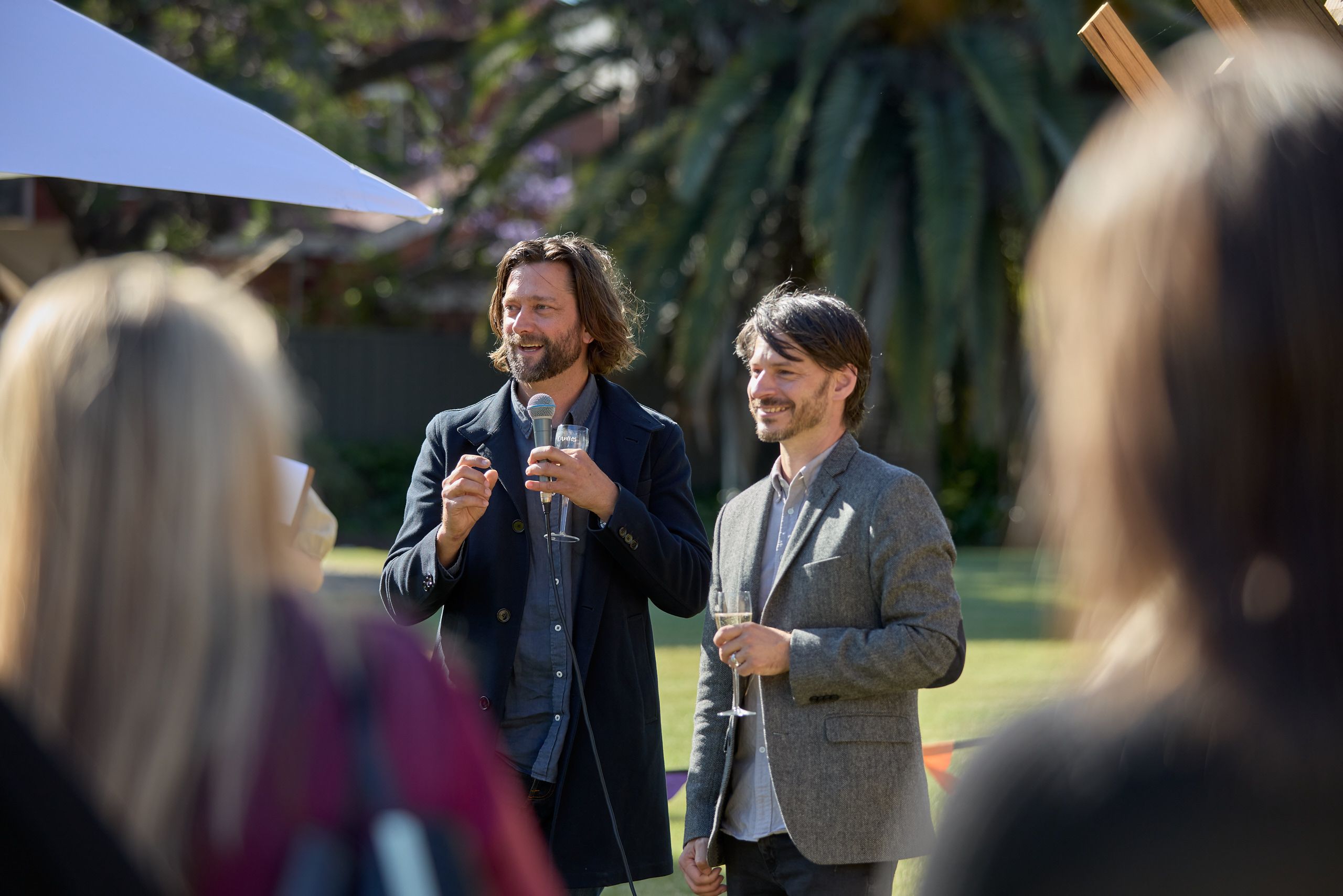 To men stand holding wine glasses in a park with a crowd gathered around, the one on the left speaks into a microphone 