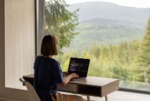 A woman in a blue t-shirt, typing on a laptop at a desk with a view of a forrest.
