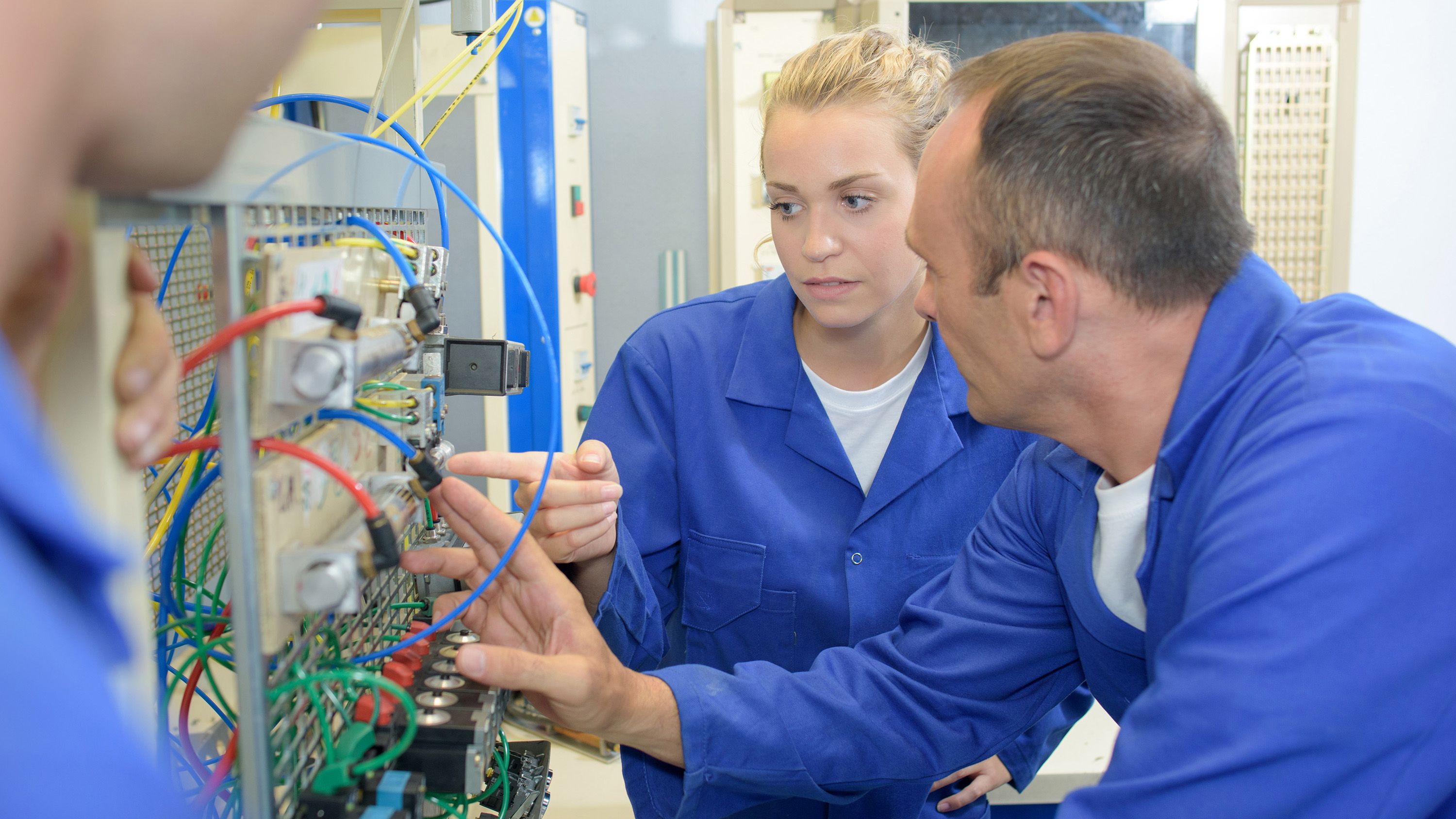 A young female worker and older male worker wearing blue overalls looking at an electrical switch board in a factory.