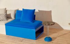 A duo-tone blue and light grey sofa with a steel base and fabric top beside a small, round side table