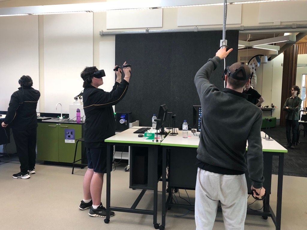 Three students stand in a classroom with VR goggles on, gesturing in the air as they play a VR game.