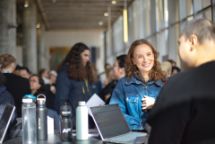 Smiling woman in a denim jacket speaks with a staff member at a busy information table during a university event.