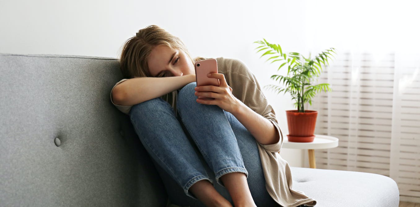 woman in jeans and a taupe shirt sitting on a grey couch in the fetal position while looking at her mobile phone