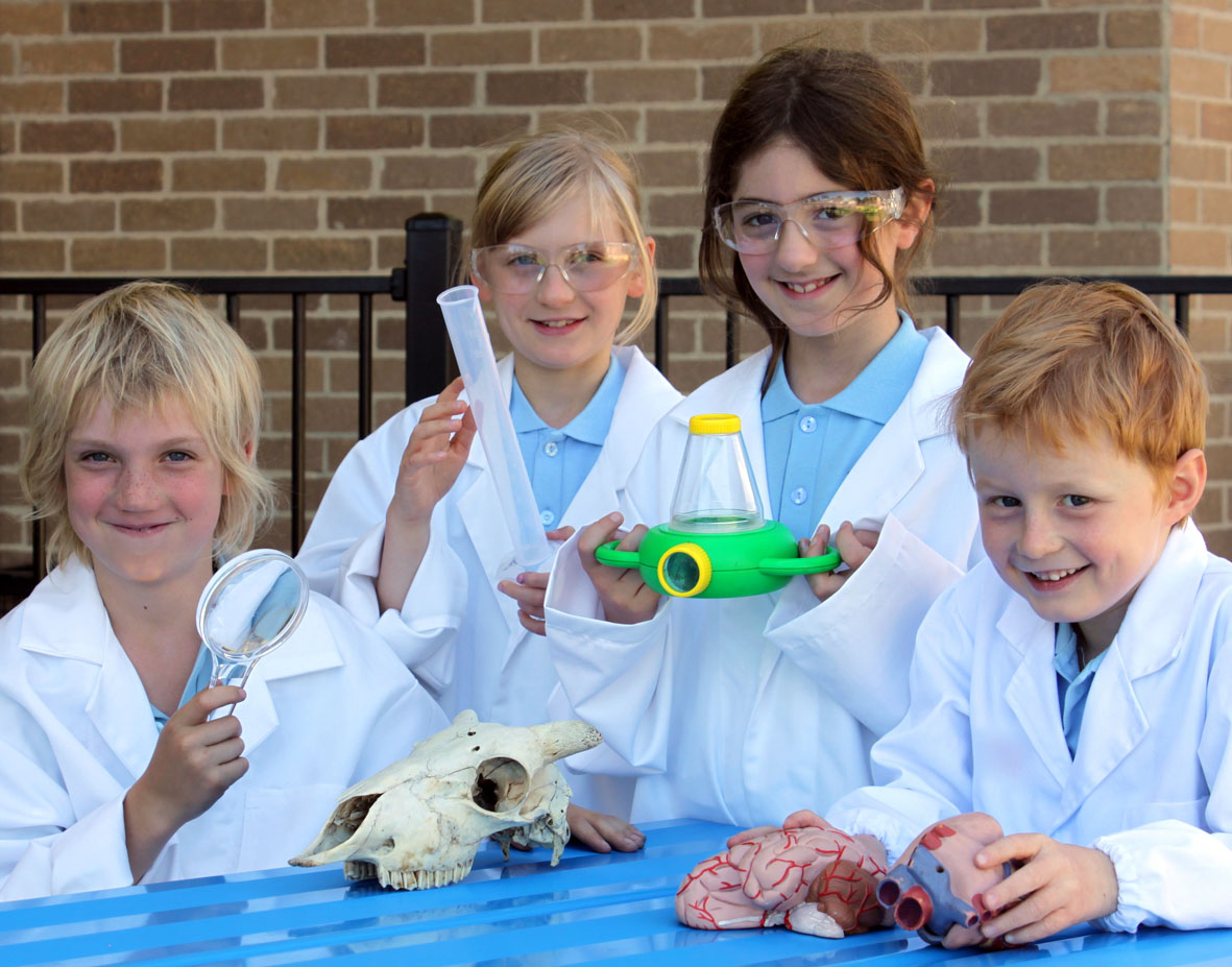 Four primary school children in white lab coats each holding a scientific object