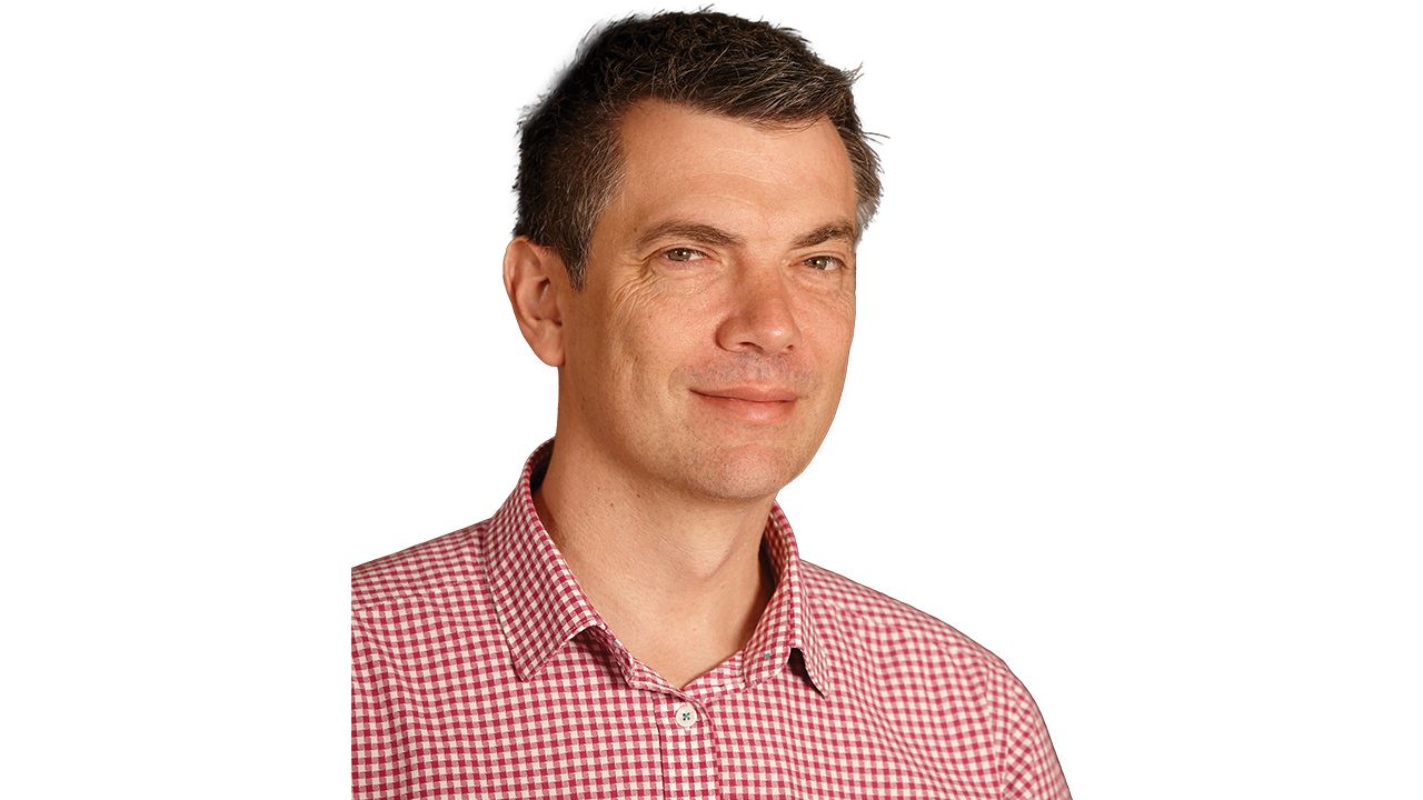 A professional portrait headshot of Will Joske wearing a red and white checkered shirt.