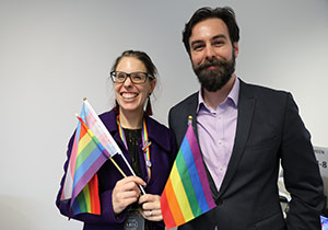 A man and a woman wear purple clothes and hold rainbow flags.