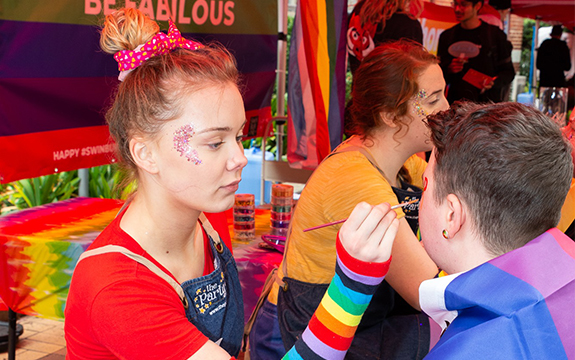 A face painting paints a student's face at the Pride Fair Day