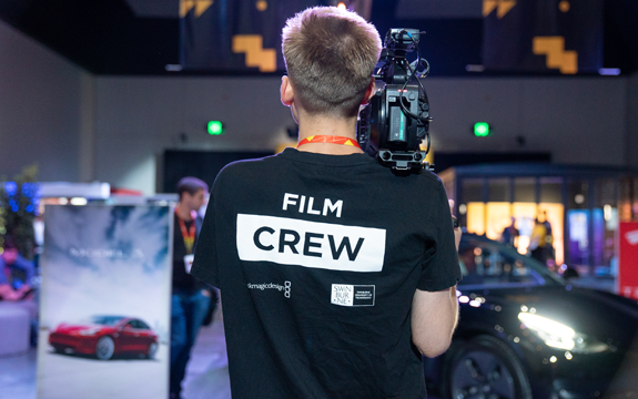 The backview of a person wearing a black tshirt that says 'Film Crew' holding a camera on their shoulder.