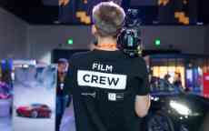 The backview of a person wearing a black tshirt that says 'Film Crew' holding a camera on their shoulder.