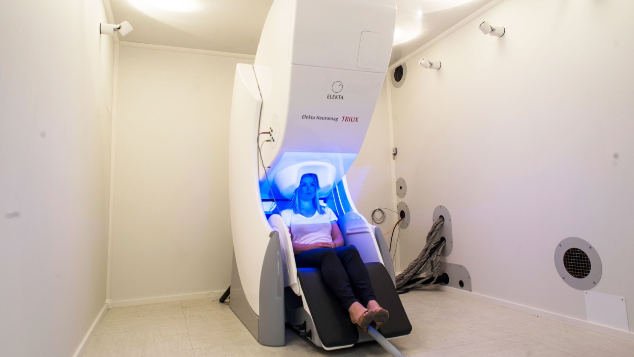 A woman sits inside a large chair like machine with her head inside a scanner. A blue light is shining around.