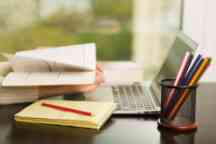 Persons hands holding a textbook at a desk with laptop, pencils and notepad.