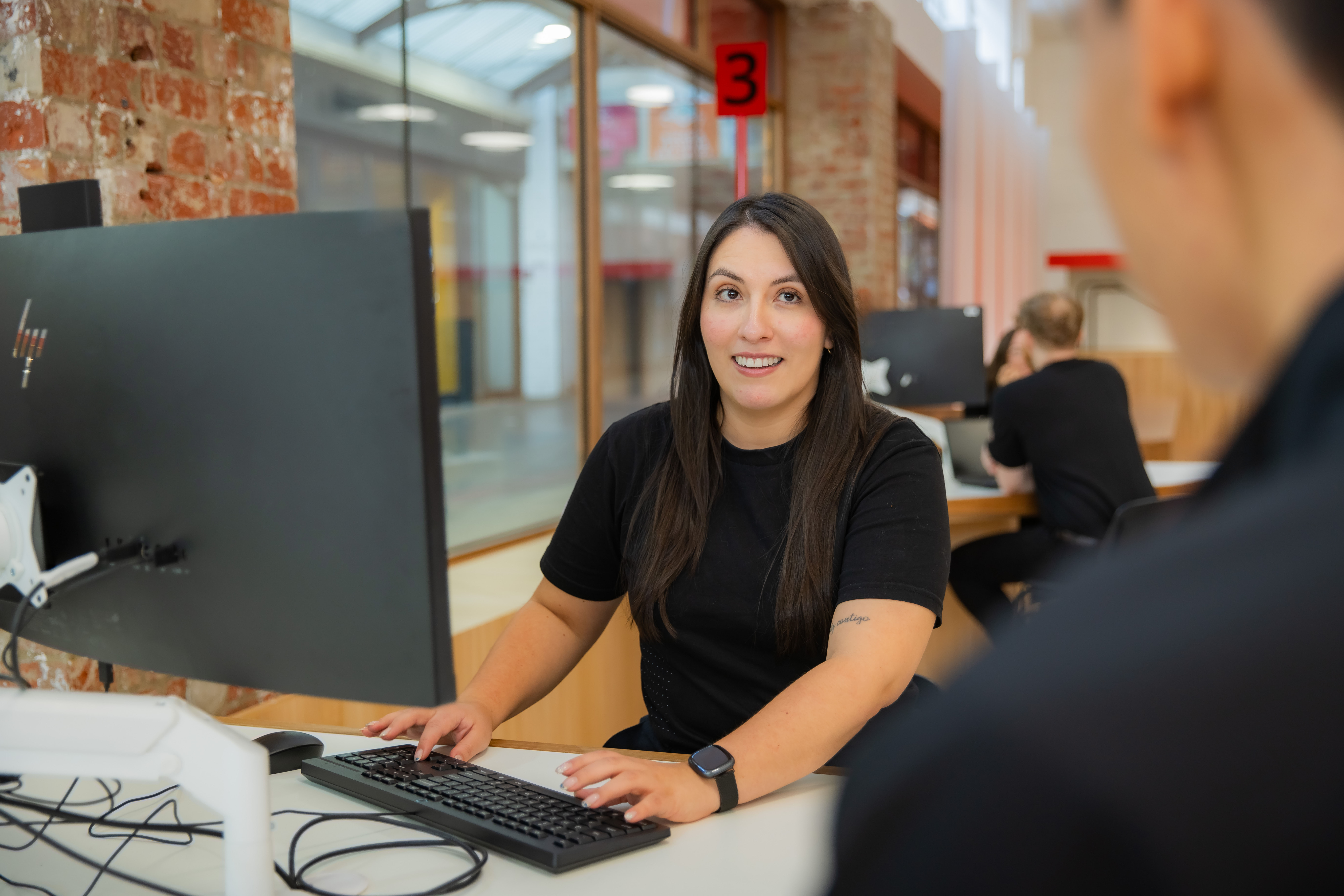 A Swinburne staff member smiles while assisting a visitor at the Future Student HQ reception desk on Hawthorn campus.