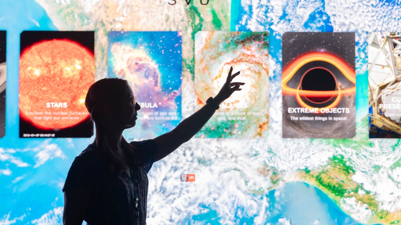 An shadowed outline of a person point to a digital screen showing stars, nebular, and other planetary things.