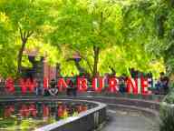 Students sitting outside by a pond around red Swinburne sign under green trees