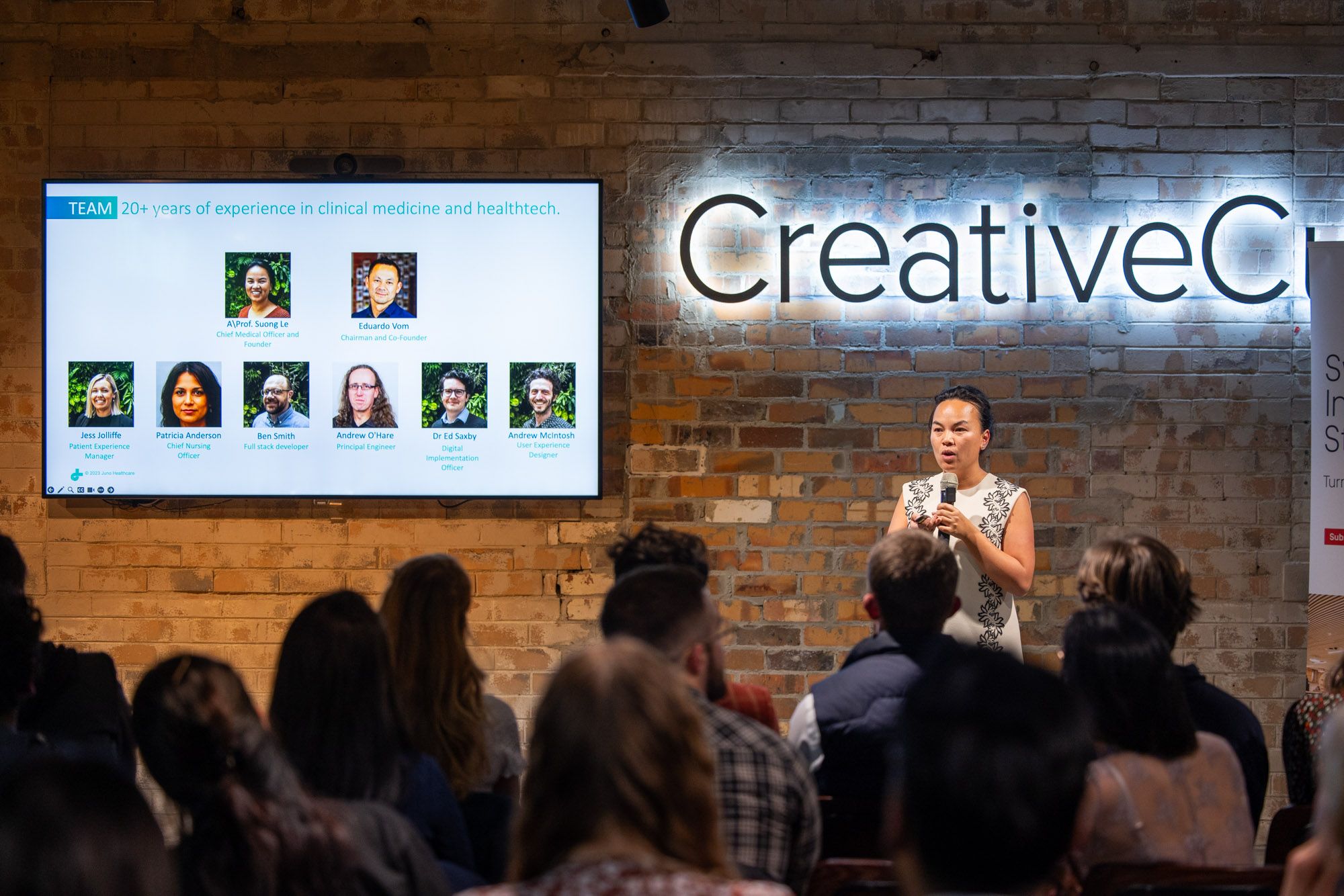 A wide shot of Suong Le, the founder of Juno Healthcare presenting at 2023 Swinburne Accelerator Program Demo Night. They're holding a microphone and are speaking to graphics visible on a display to their side. The shadow of the audience is visible in the foreground. 