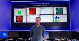 Andrew Jackling at the Australian Space Weather Forecasting centre standing infront of his work station with various monitors showing space weather data
