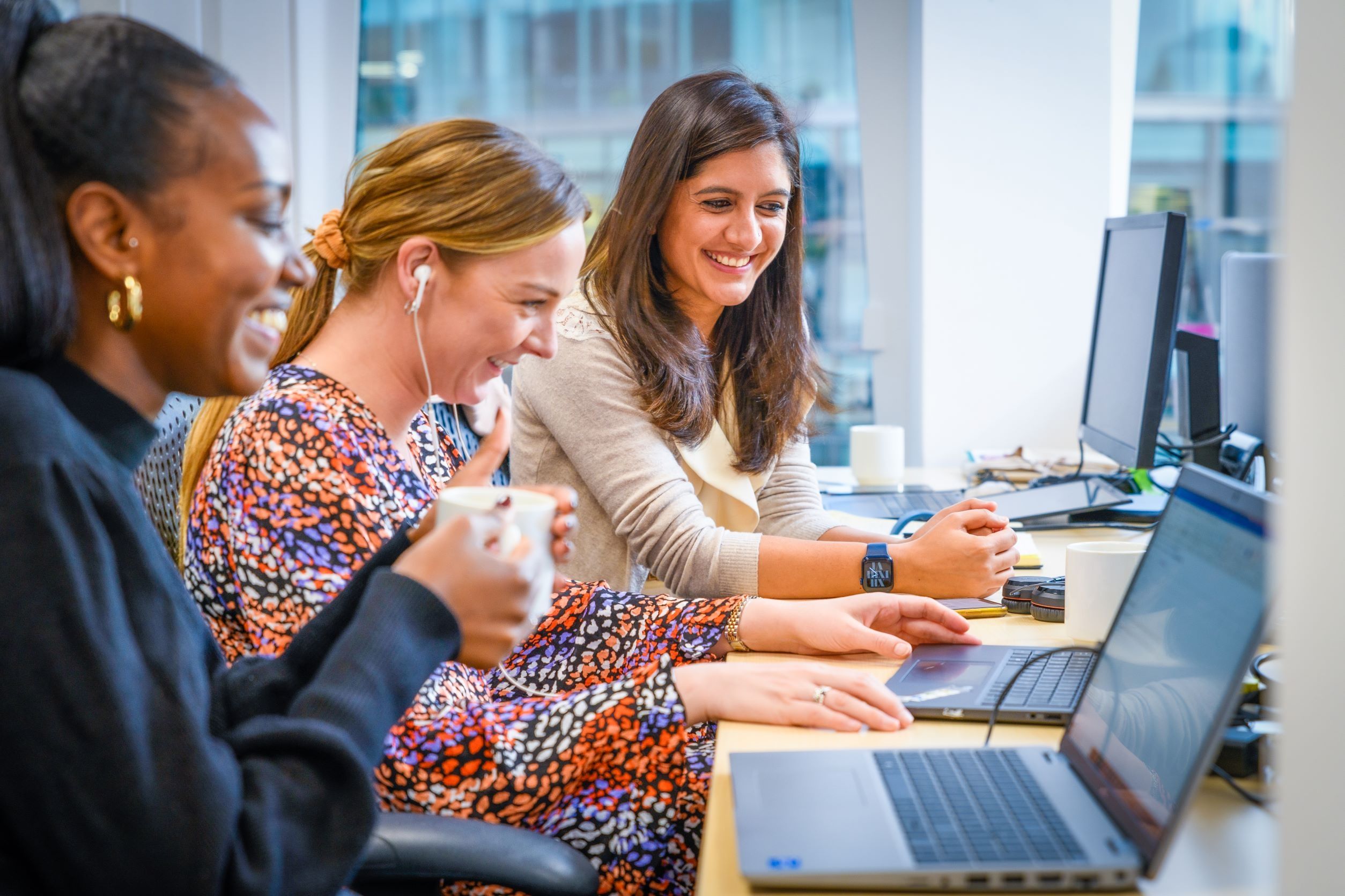 three women sitting at a desk smiling while they work on their laptops