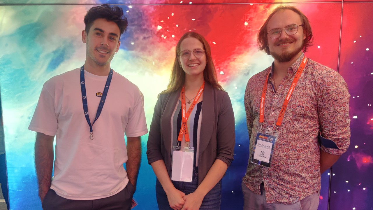 Swinburne PhD students, featuring Daniel Ricardo, Belinda Rich and Philipp Zanon stand side by side in front of a bright galaxy like backdrop