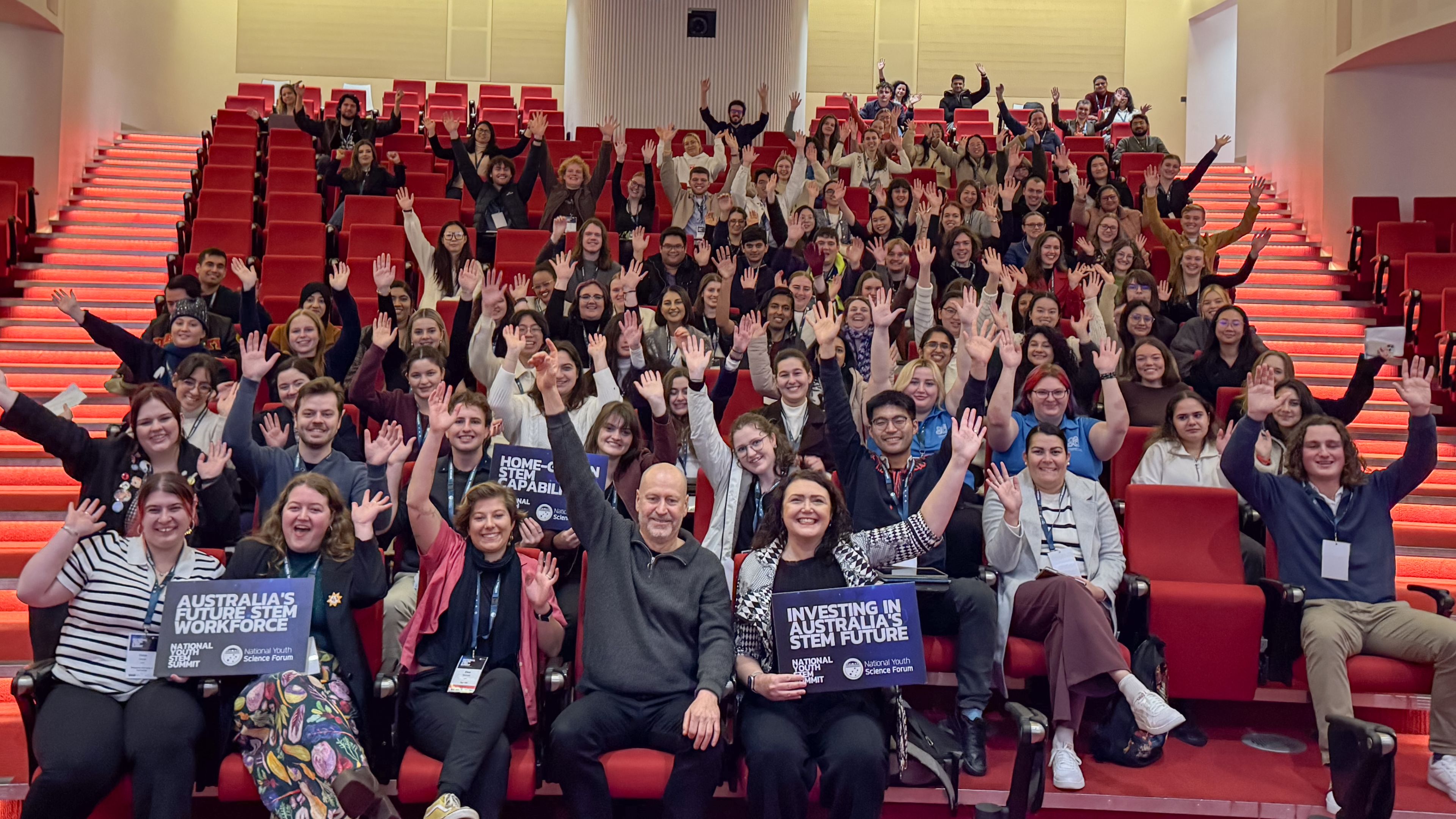 Attendees of a summit in a lecture theatre holding up their arms in the air.
