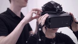 A person wearing black shirt adjusts the virtual reality headset worn by second person, who is also wearing a black bodysuit featuring small white balls which are motion sensors