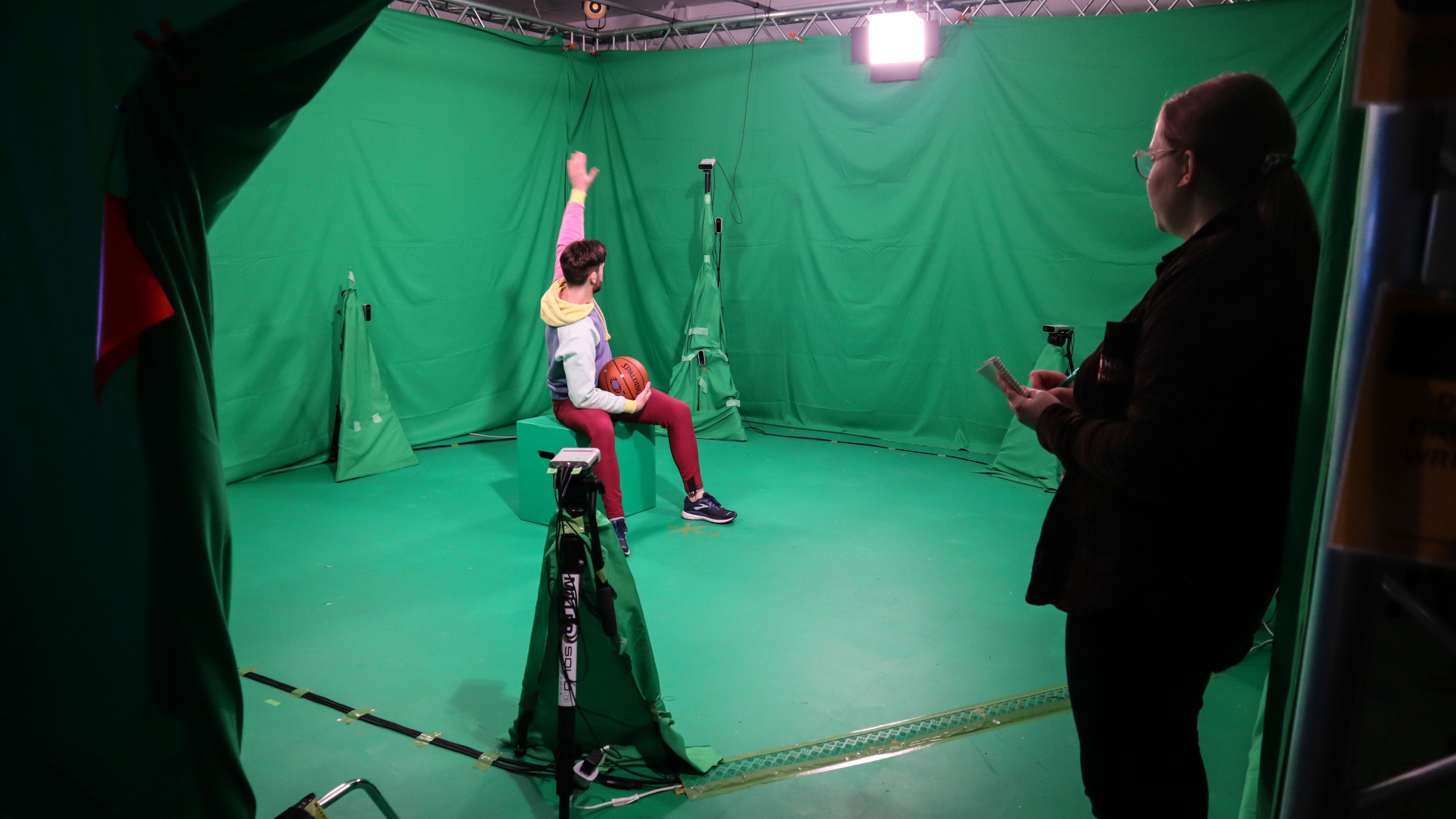 A person shits on a stool in a green studio surrounded by by cameras 